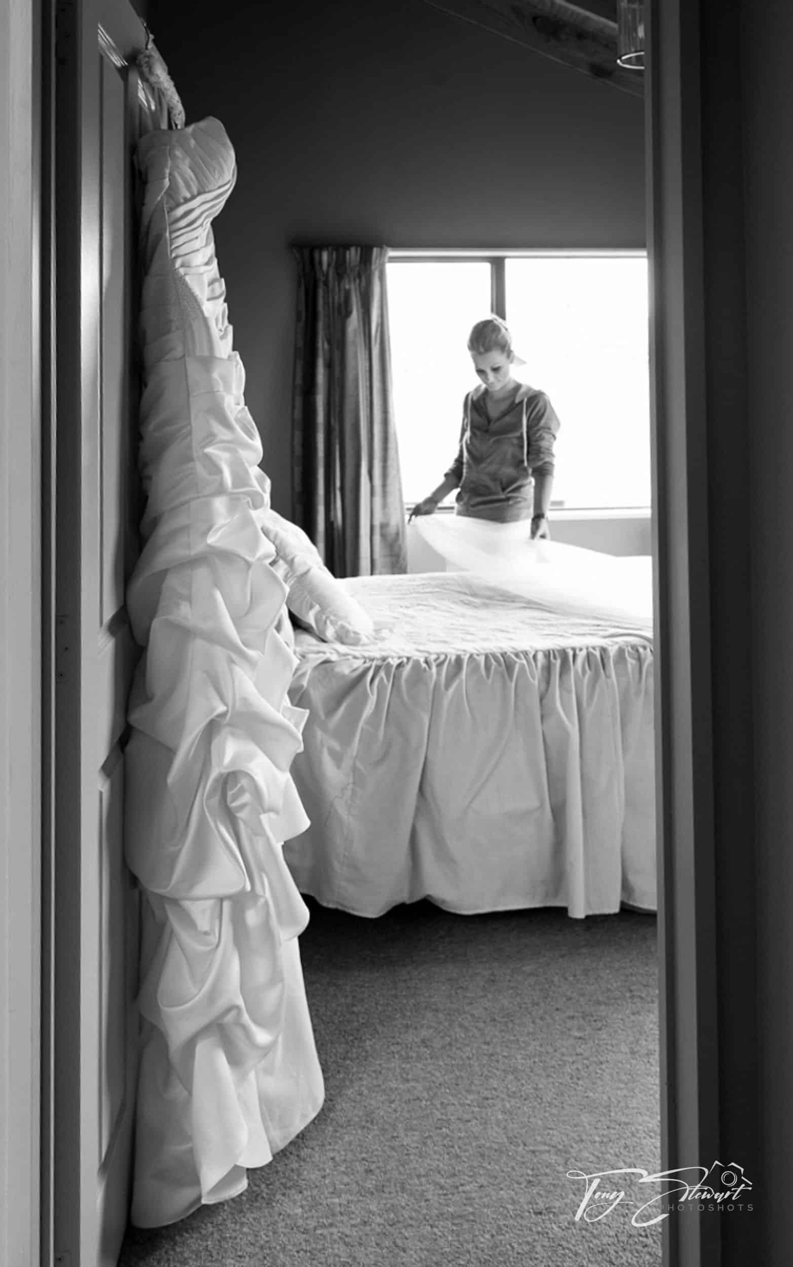 Bride prepares her veil in a bedroom while her dress is hung on the door.