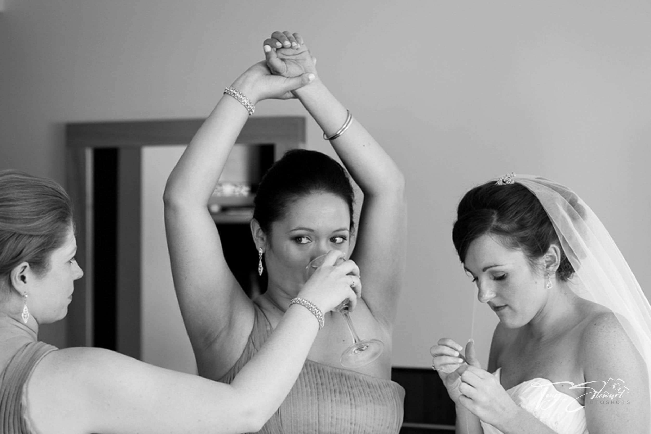Bridesmaids receives a glass of wine, while holding up her arm for the bride to help sew her dress.