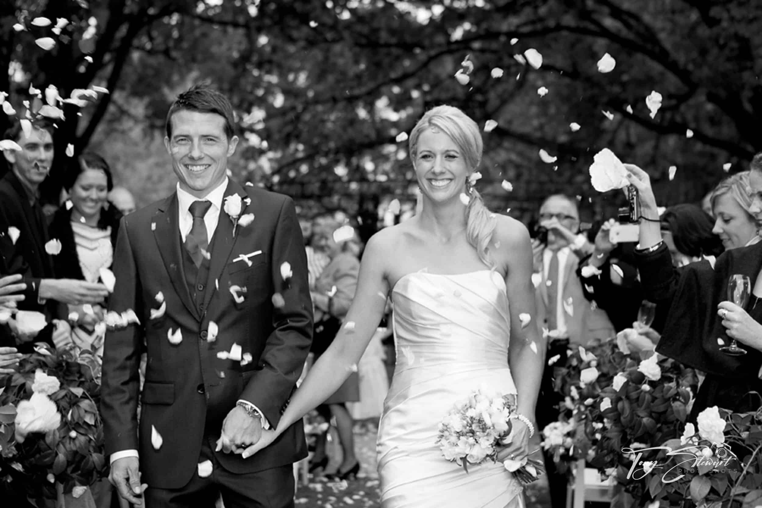 Happy couple leave their wedding ceremony walking down aisle showered with petals.