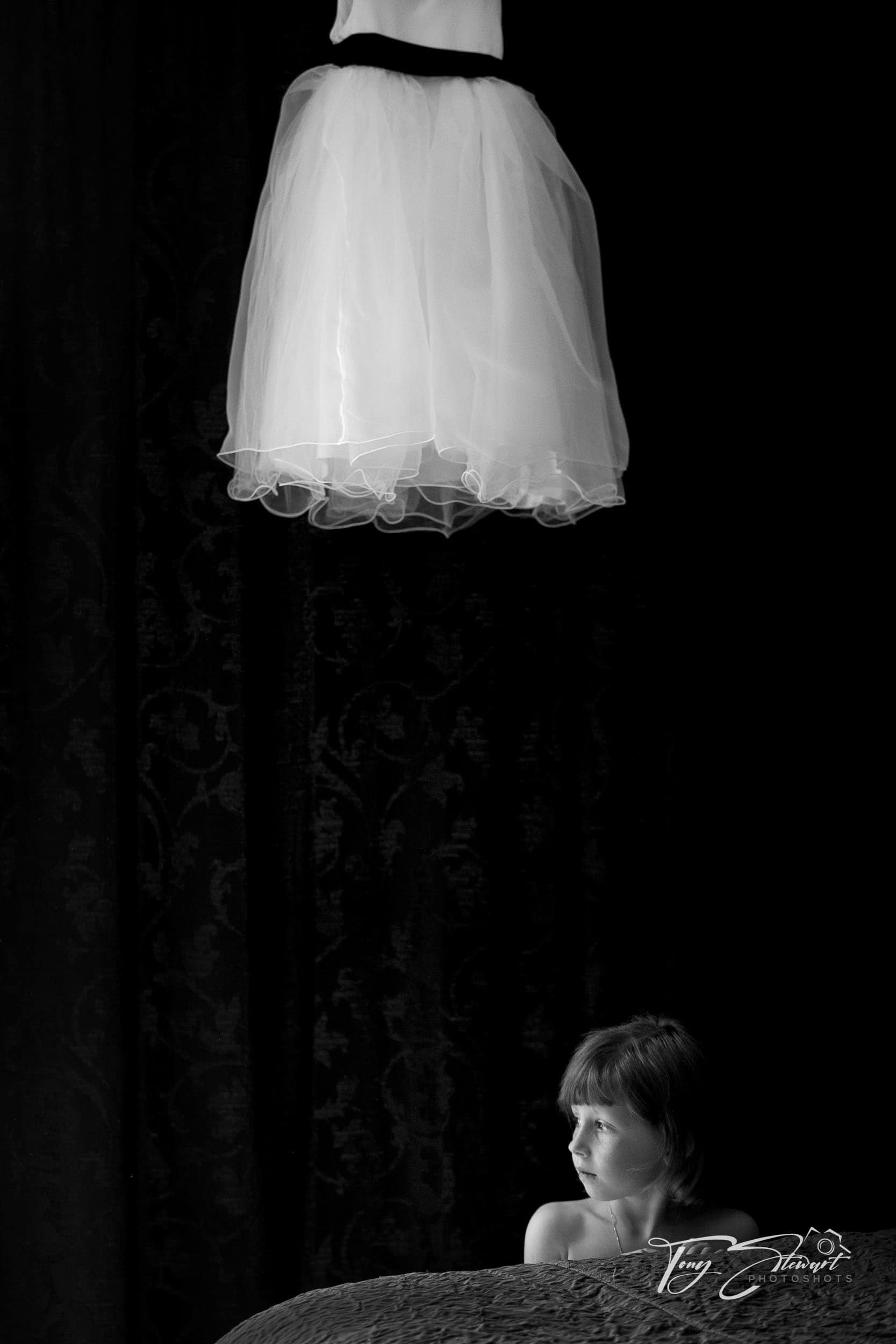 A young bridesmaid poses whistfully by a bed under large wedding dress, lit by strong side window light.