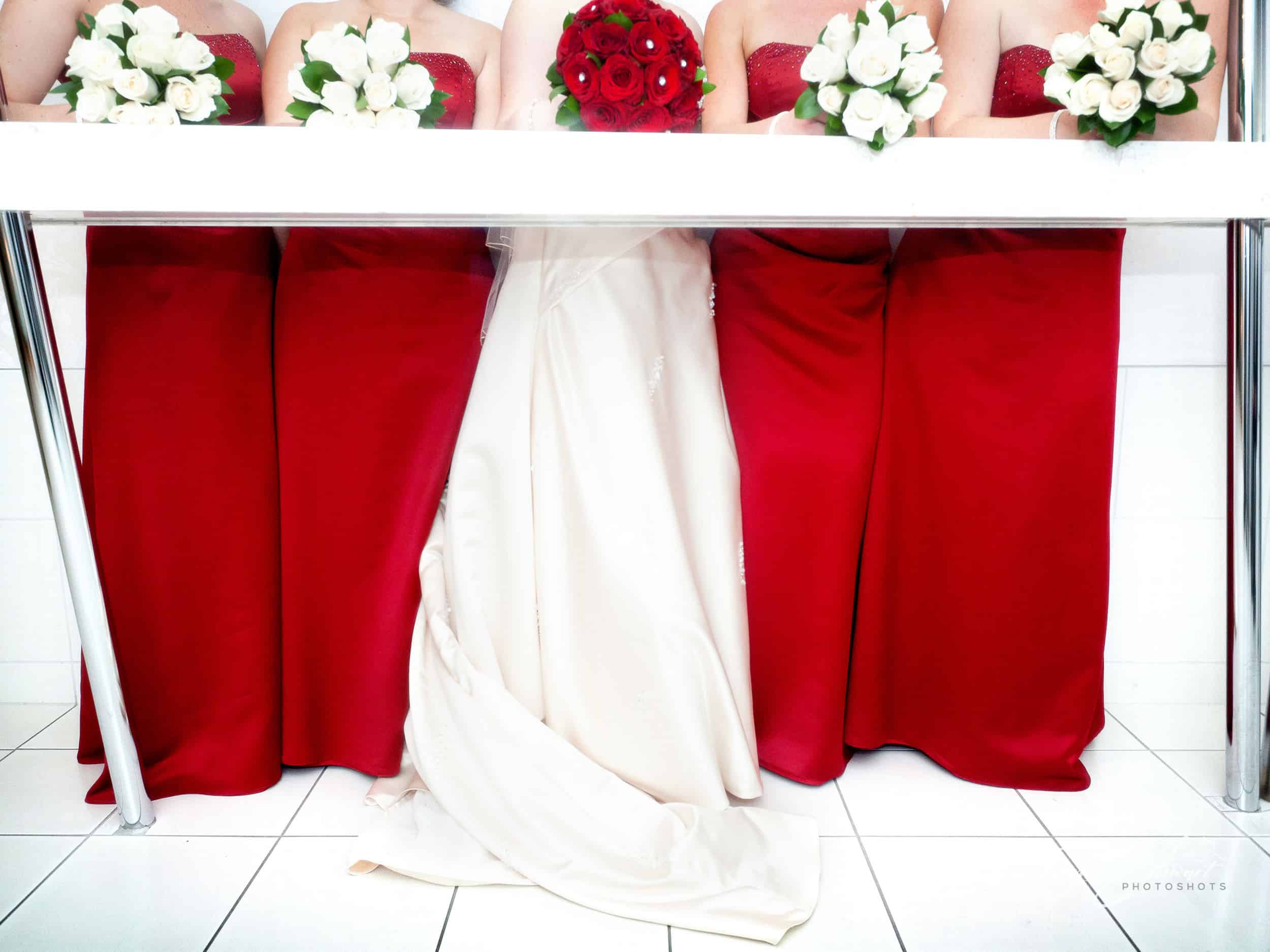 Colourful bridesmaids in red flag a bride in white, while leaning on a table with their heads obscured, at the George hotel, Christchurch.