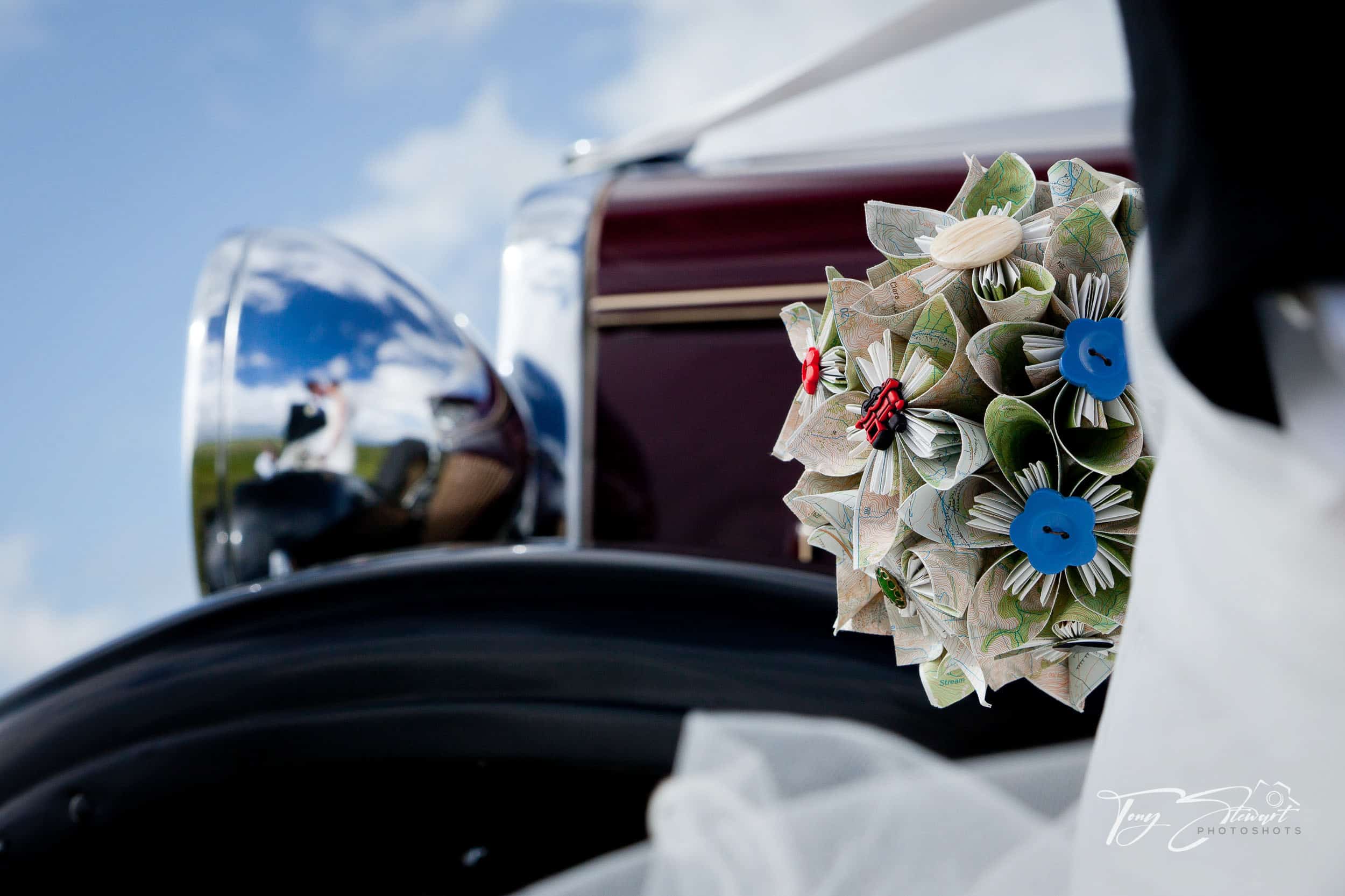 Bridal couple partly obscured seen in reflection of vintage car headlight holding holding a bouquet of paper flowers.