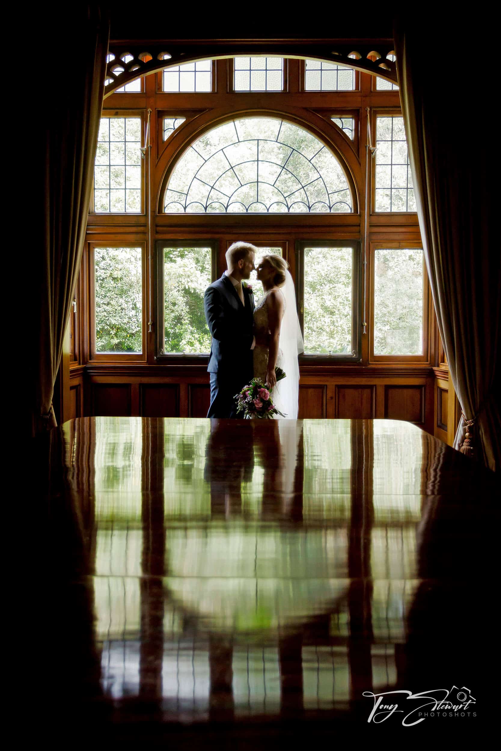 Bridal couple stand side on in rfont of a grand arched window at Otahuna Lodge.