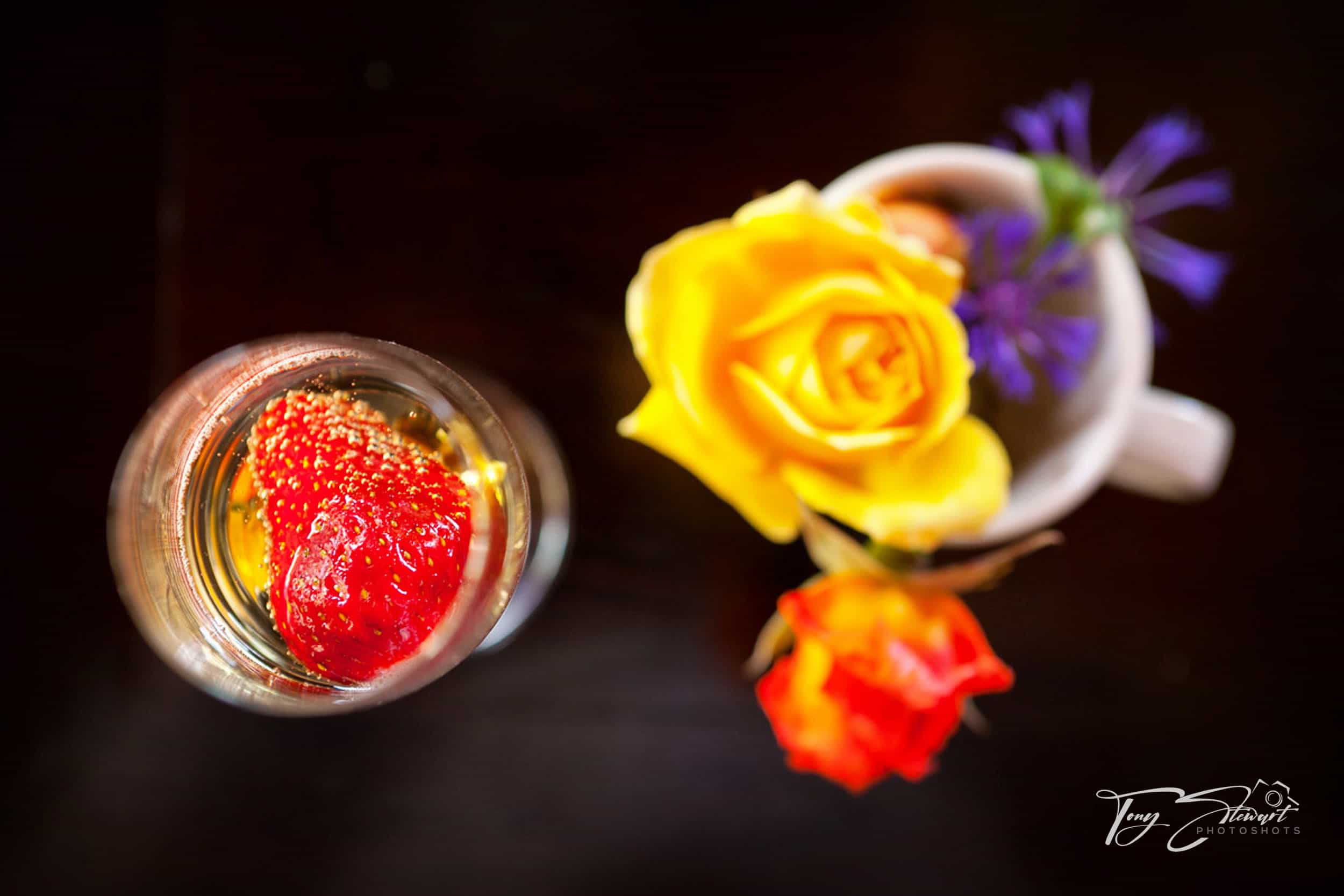 Strawberry in wine glass stands beside a posy of bright flowers in a mug viewed from above.