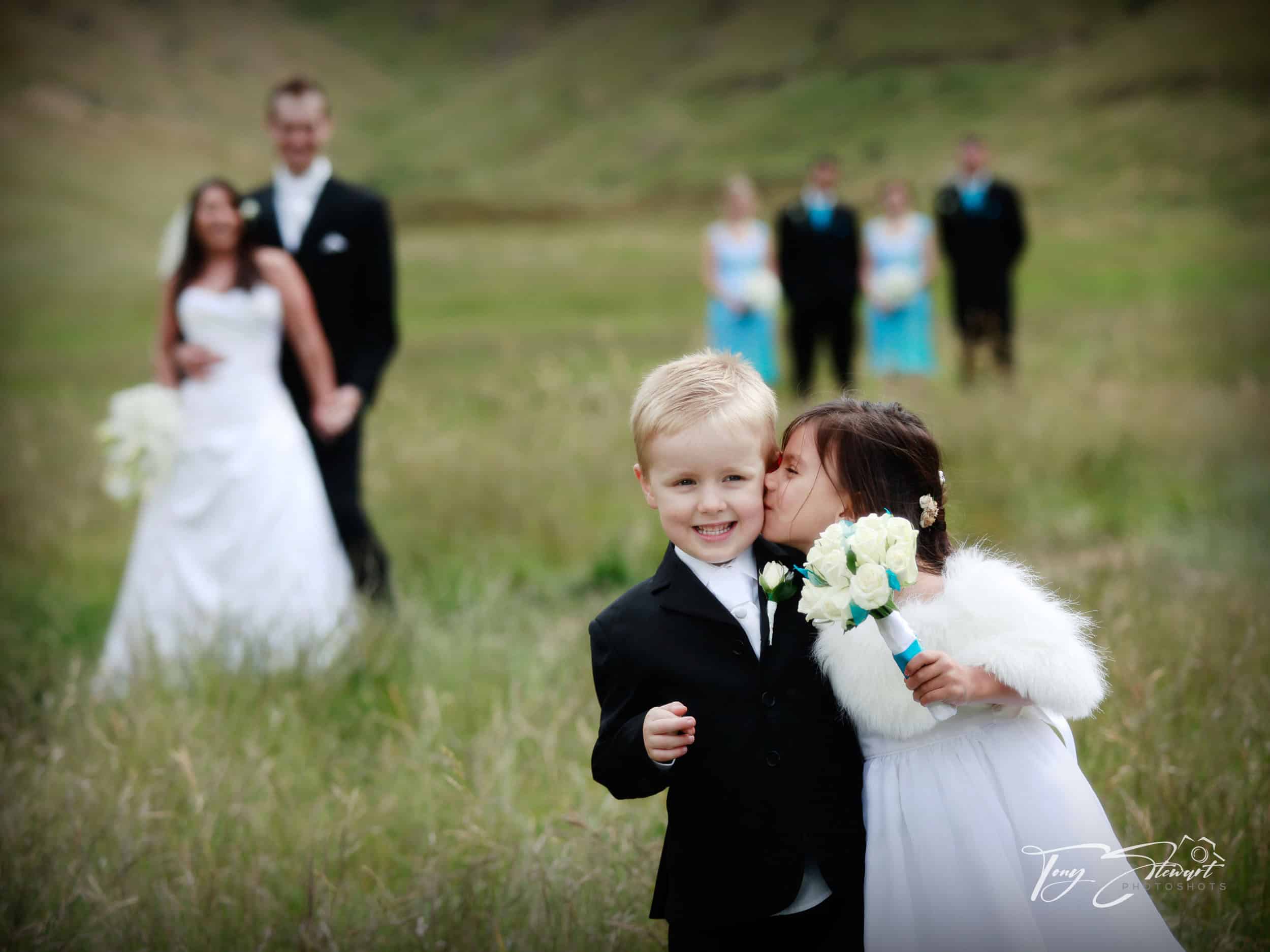 Cute page boy and girl replicate earlier kiss of bridal couple standing in a field.