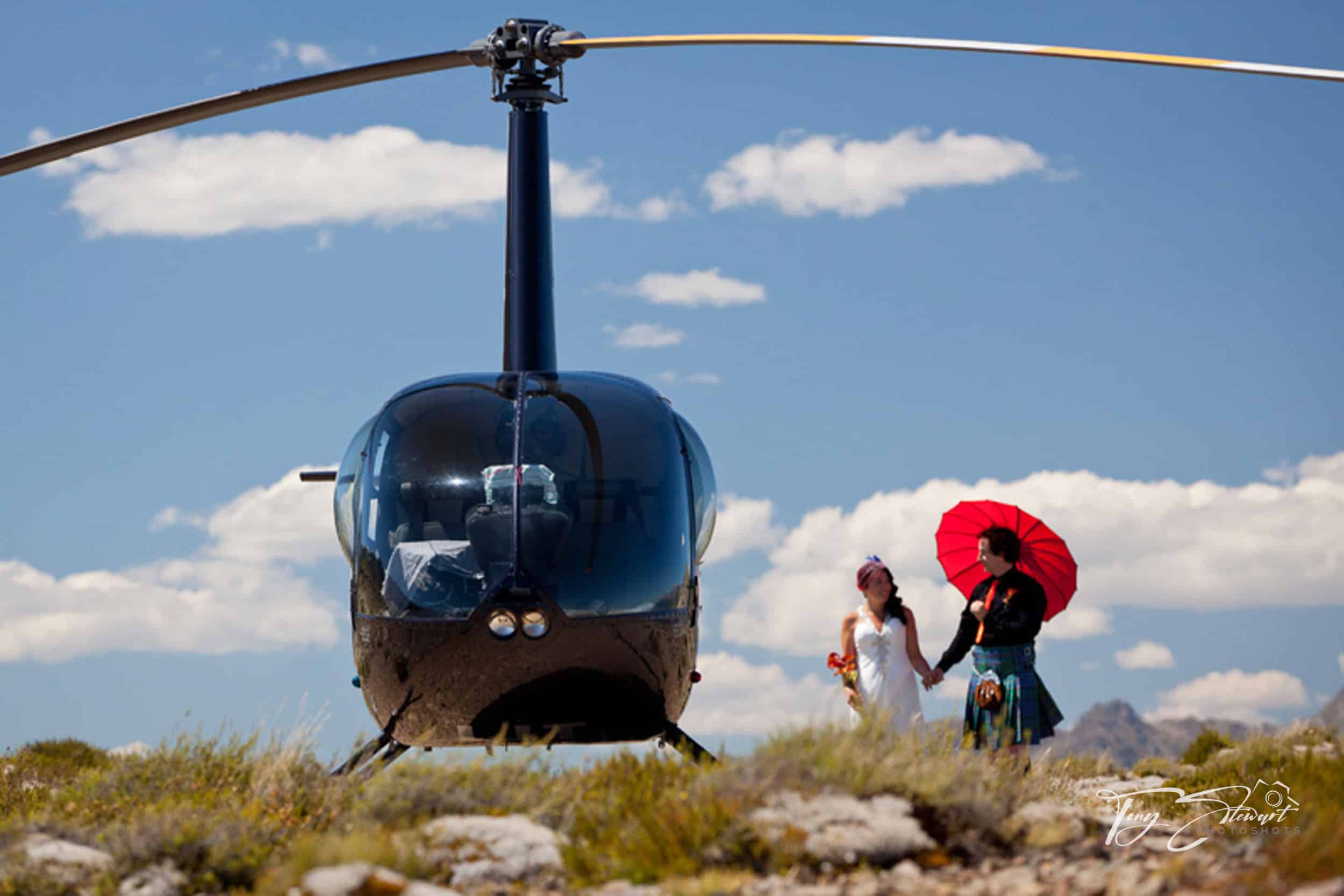 Bride and groom walk on a hilltop towards their helicopter while holding bright red parasol.